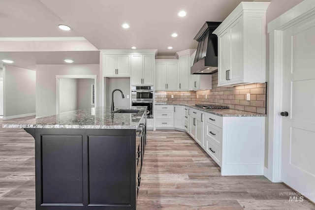 kitchen featuring light stone countertops, light hardwood / wood-style floors, wall chimney range hood, and a center island with sink