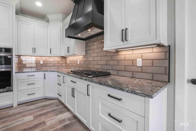 kitchen featuring light wood-type flooring, stainless steel appliances, dark stone counters, white cabinets, and custom range hood