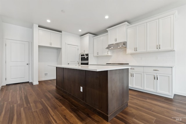 kitchen featuring tasteful backsplash, dark wood-type flooring, under cabinet range hood, an island with sink, and white cabinetry