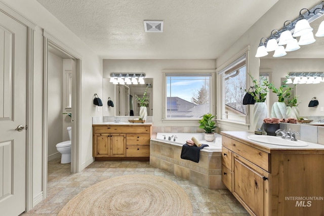 bathroom featuring toilet, two vanities, a bath, a textured ceiling, and a sink