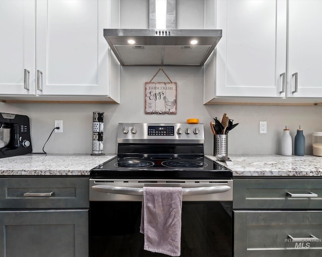 kitchen with stainless steel electric stove, wall chimney range hood, light stone counters, and white cabinetry