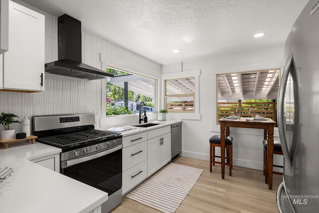 kitchen with appliances with stainless steel finishes, wall chimney exhaust hood, a sink, light wood-style flooring, and a textured ceiling