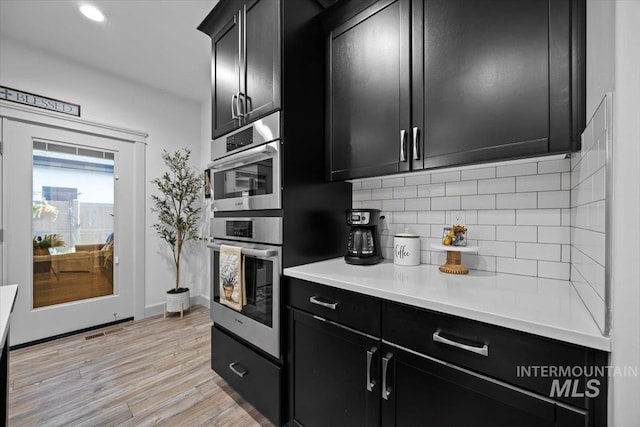 kitchen with double oven, light wood-style floors, light countertops, dark cabinetry, and backsplash