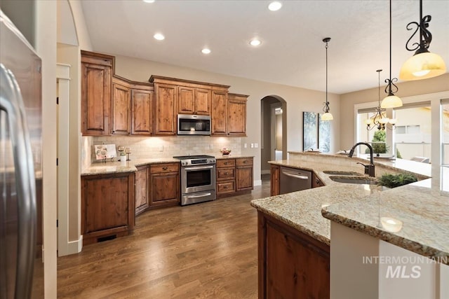 kitchen featuring brown cabinetry, decorative backsplash, stainless steel appliances, arched walkways, and a sink