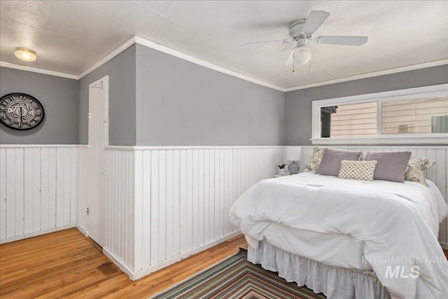 bedroom with a wainscoted wall, ornamental molding, a ceiling fan, and light wood finished floors