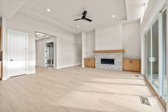 unfurnished living room featuring ceiling fan, a tray ceiling, light hardwood / wood-style flooring, and a stone fireplace