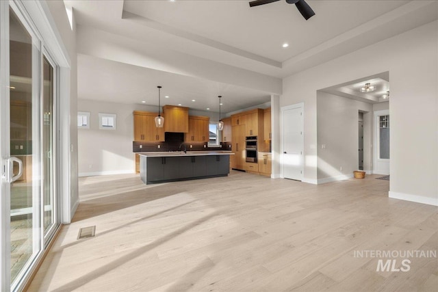 kitchen with double oven, an island with sink, hanging light fixtures, a raised ceiling, and ceiling fan