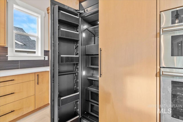 kitchen with light wood-type flooring, stainless steel double oven, backsplash, and light brown cabinets