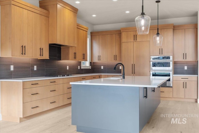 kitchen featuring double oven, light brown cabinetry, decorative backsplash, hanging light fixtures, and a kitchen island with sink