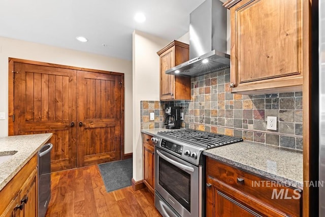 kitchen with appliances with stainless steel finishes, wall chimney range hood, dark wood-style flooring, decorative backsplash, and light stone counters