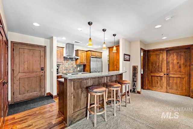 kitchen featuring stainless steel refrigerator, wall chimney range hood, brown cabinetry, a breakfast bar area, and a peninsula