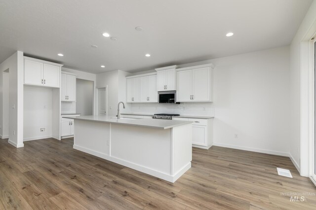 kitchen with stainless steel microwave, tasteful backsplash, white cabinetry, an island with sink, and light wood-style floors