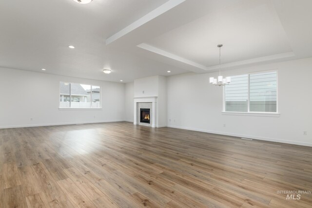 unfurnished living room featuring a warm lit fireplace, light wood-style flooring, baseboards, a raised ceiling, and an inviting chandelier