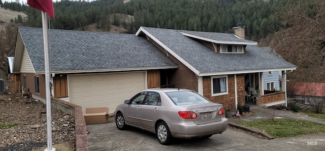 view of front of home with concrete driveway, a view of trees, roof with shingles, and a chimney