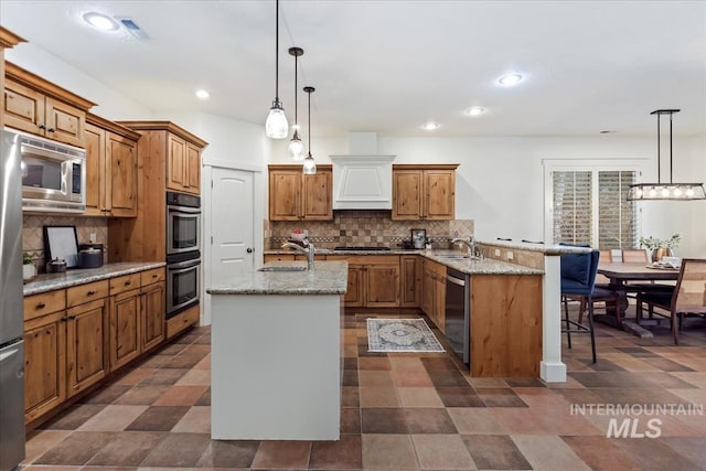 kitchen with an island with sink, a peninsula, brown cabinetry, stainless steel appliances, and a sink