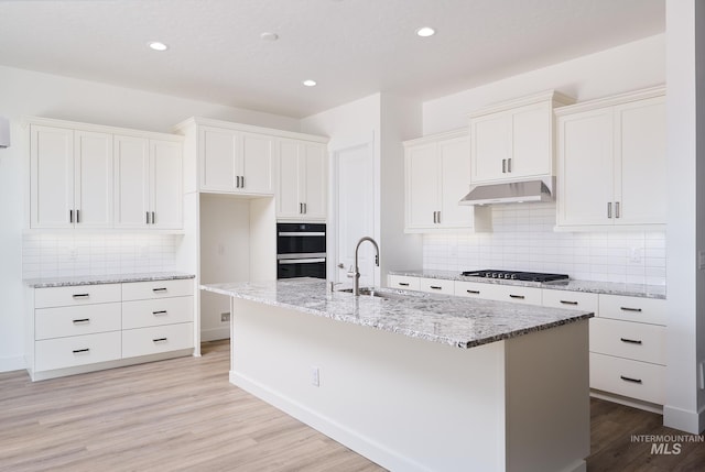 kitchen featuring light wood-type flooring, white cabinetry, under cabinet range hood, a sink, and a center island with sink