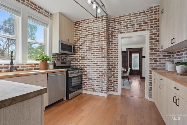 kitchen with light brown cabinetry, appliances with stainless steel finishes, light wood-style floors, brick wall, and butcher block countertops