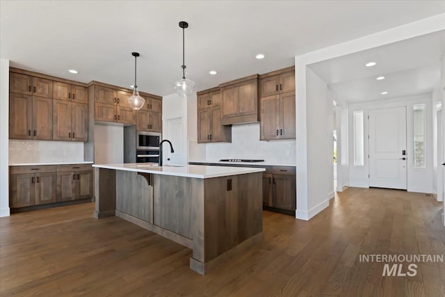 kitchen featuring sink, dark wood-type flooring, stainless steel appliances, pendant lighting, and a kitchen island with sink