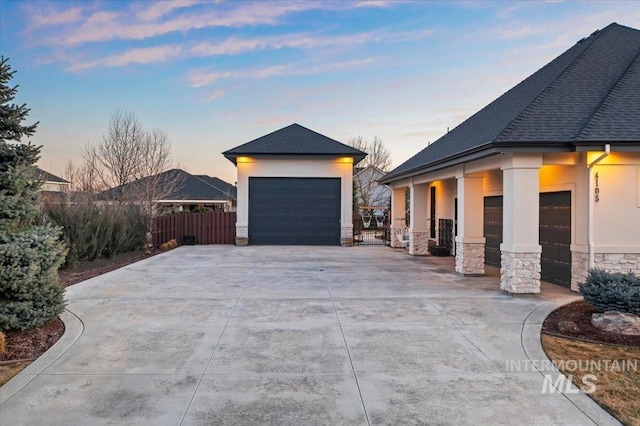 property exterior at dusk with a garage, fence, stone siding, roof with shingles, and stucco siding