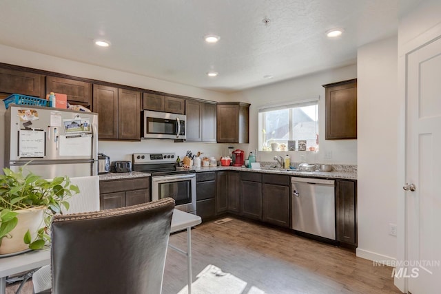 kitchen with light wood-type flooring, recessed lighting, stainless steel appliances, light stone countertops, and dark brown cabinets