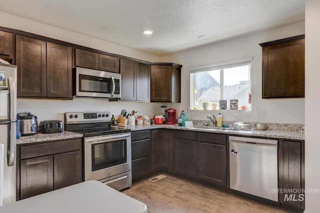 kitchen with dark brown cabinetry, light stone counters, light wood-style floors, stainless steel appliances, and a sink