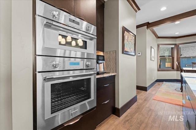 kitchen with dark brown cabinets, stainless steel double oven, ornamental molding, and light wood-type flooring