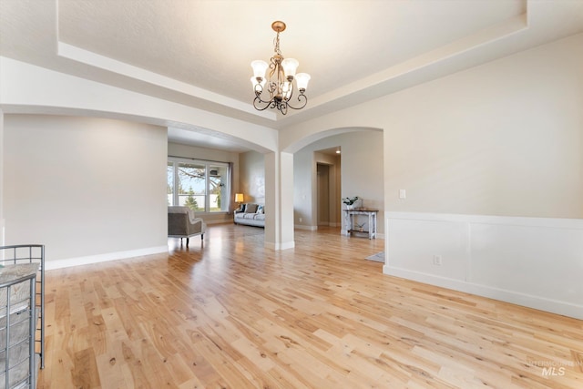 interior space featuring arched walkways, a raised ceiling, a wainscoted wall, an inviting chandelier, and light wood-style floors