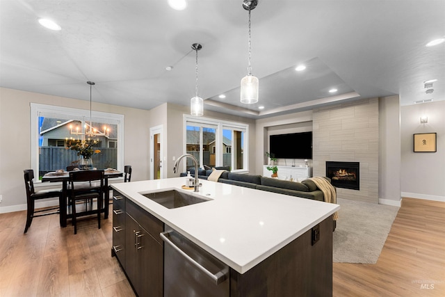 kitchen featuring a center island with sink, sink, stainless steel dishwasher, dark brown cabinetry, and a tiled fireplace