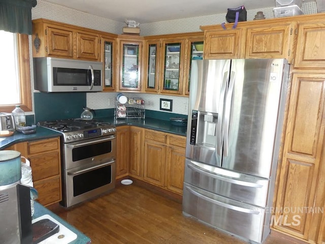 kitchen with wallpapered walls, dark wood finished floors, brown cabinetry, glass insert cabinets, and stainless steel appliances