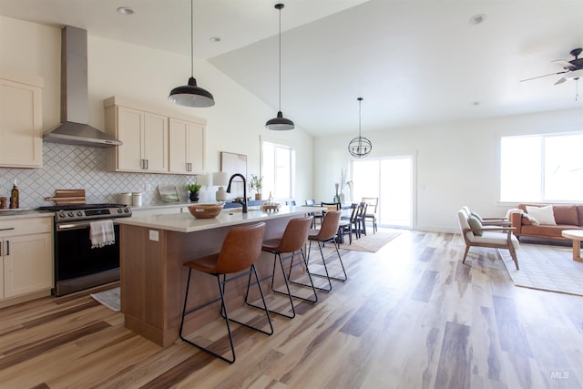 kitchen featuring wall chimney range hood, stainless steel range with electric cooktop, light wood-style floors, a kitchen island with sink, and light countertops