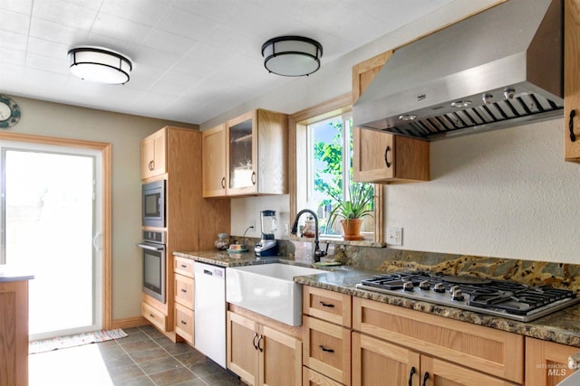 kitchen featuring light brown cabinets, wall chimney exhaust hood, dark stone counters, appliances with stainless steel finishes, and a sink