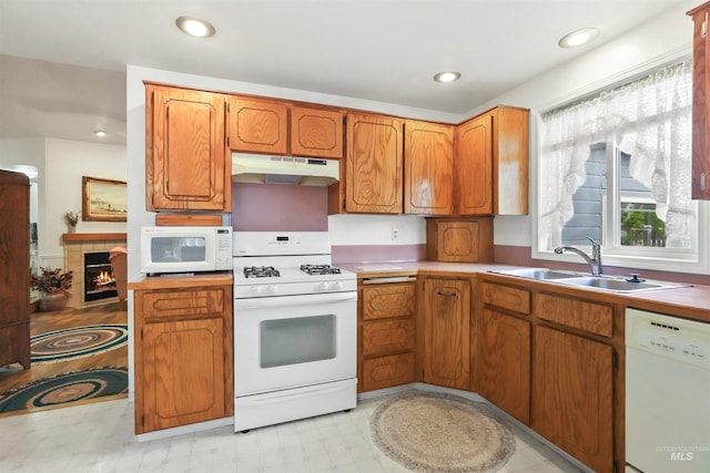 kitchen with brown cabinetry, a sink, light floors, under cabinet range hood, and white appliances