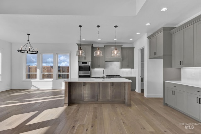 kitchen featuring stainless steel double oven, a center island with sink, gray cabinets, light hardwood / wood-style floors, and hanging light fixtures