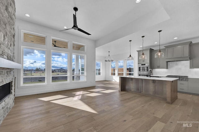 kitchen featuring ceiling fan with notable chandelier, a stone fireplace, hardwood / wood-style flooring, an island with sink, and tasteful backsplash