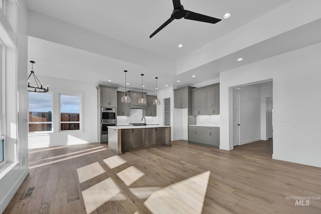 kitchen with light wood-type flooring, stainless steel double oven, sink, pendant lighting, and an island with sink