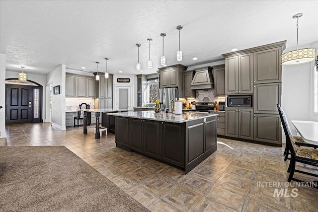 kitchen with light stone counters, a center island with sink, custom range hood, backsplash, and appliances with stainless steel finishes