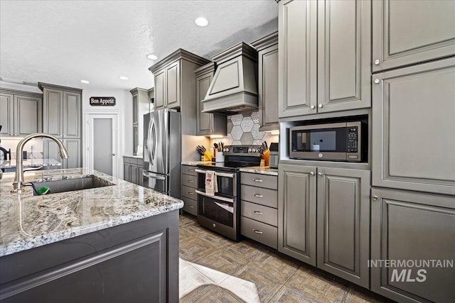 kitchen with stainless steel appliances, custom range hood, backsplash, a sink, and light stone countertops