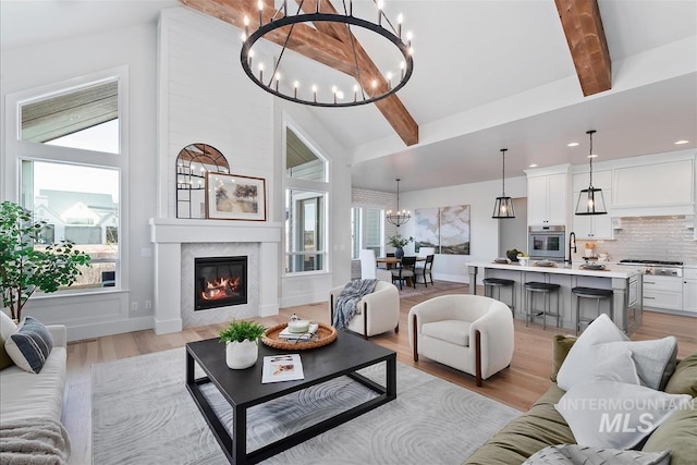 living room with a chandelier, beamed ceiling, a glass covered fireplace, and light wood-style flooring