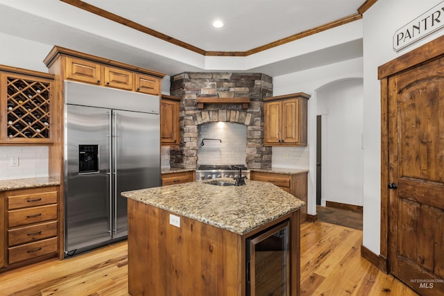 kitchen with light stone counters, built in refrigerator, a center island with sink, beverage cooler, and backsplash