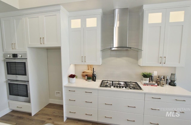 kitchen with appliances with stainless steel finishes, tasteful backsplash, hardwood / wood-style flooring, white cabinetry, and wall chimney exhaust hood