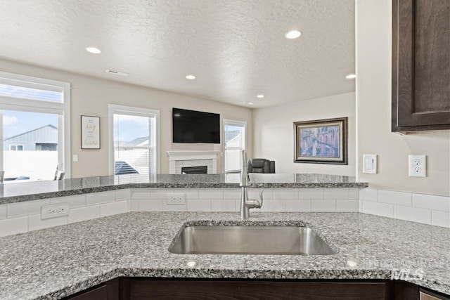kitchen with dark brown cabinetry, open floor plan, light stone counters, a textured ceiling, and a sink