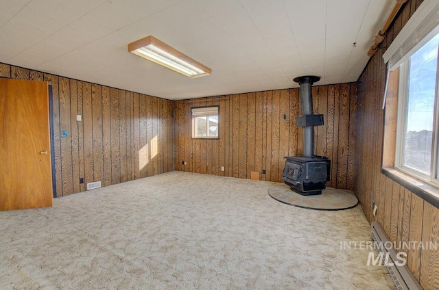 carpeted empty room featuring plenty of natural light, wood walls, a wood stove, and baseboard heating