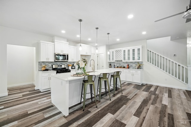 kitchen featuring a kitchen island with sink, a sink, wood finished floors, appliances with stainless steel finishes, and glass insert cabinets