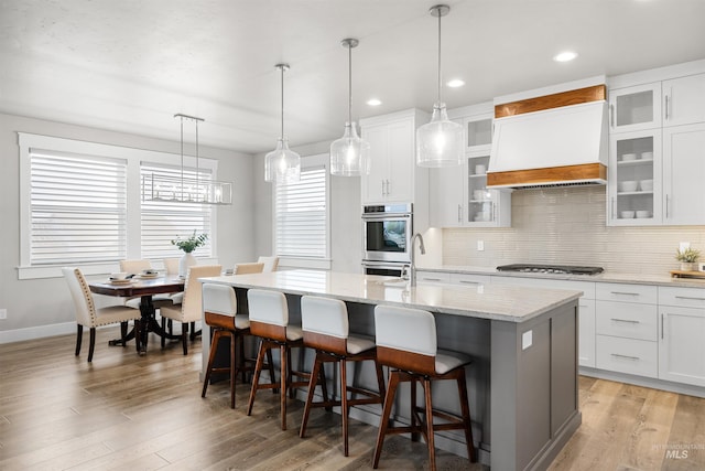 kitchen with stainless steel double oven, gas stovetop, decorative backsplash, an island with sink, and custom exhaust hood