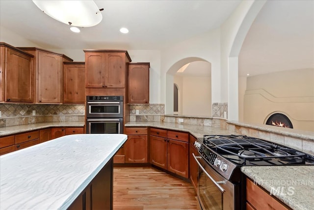 kitchen with double wall oven, light wood-style flooring, stainless steel range with gas stovetop, arched walkways, and tasteful backsplash