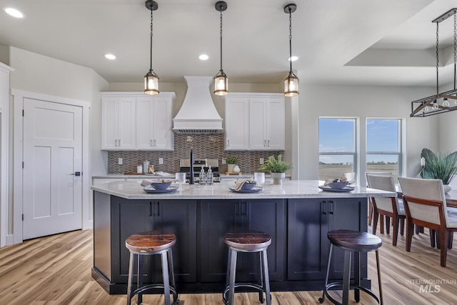 kitchen with premium range hood, tasteful backsplash, a kitchen island with sink, light wood-style flooring, and white cabinetry