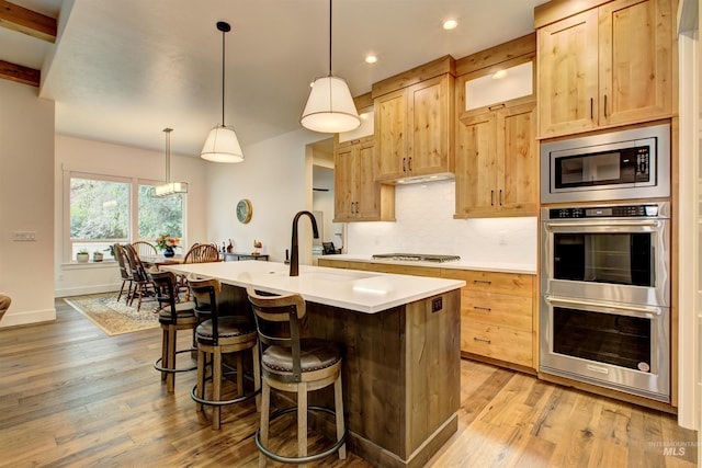 kitchen featuring pendant lighting, stainless steel appliances, an island with sink, and light brown cabinets