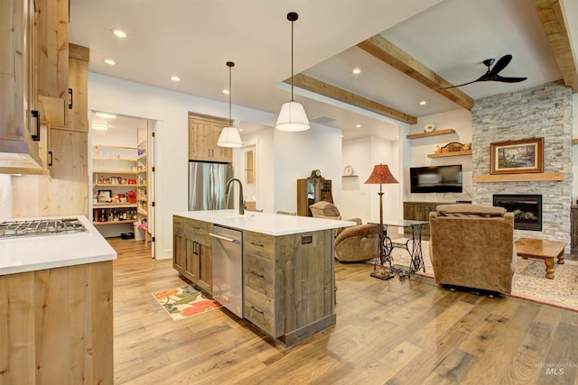 kitchen featuring a stone fireplace, hanging light fixtures, stainless steel appliances, a center island with sink, and light wood-type flooring