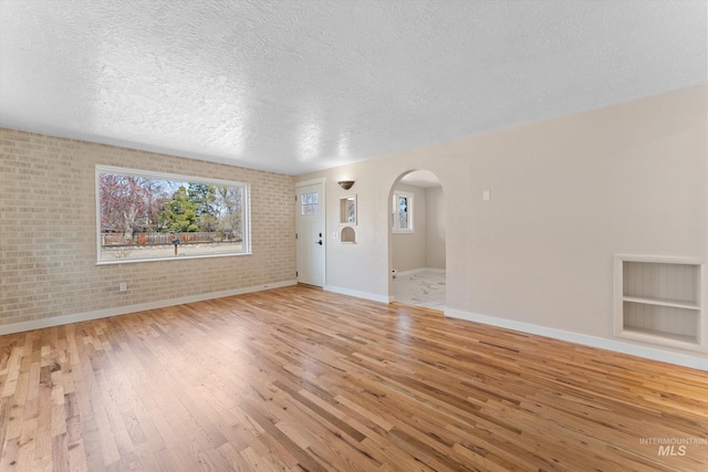 spare room featuring light wood-style flooring, a textured ceiling, arched walkways, brick wall, and baseboards