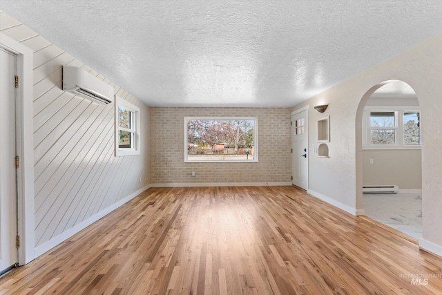 unfurnished living room featuring an AC wall unit, light wood-type flooring, baseboard heating, arched walkways, and a textured ceiling